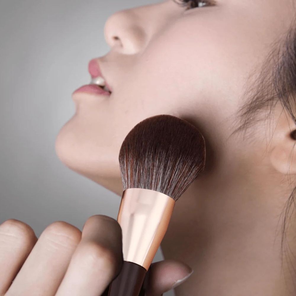 Makeup brush being used on a person's face against a neutral background
