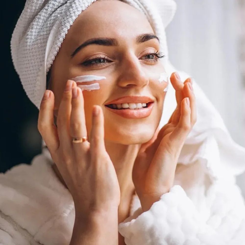 Woman applying cream to her face with a towel on her head, wearing a robe.